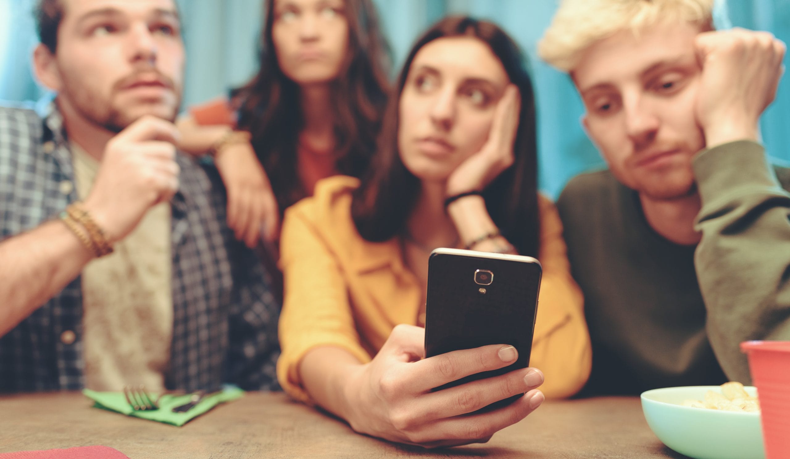 Group of friends thinking and feeling bored at home, one is holding a smartphone