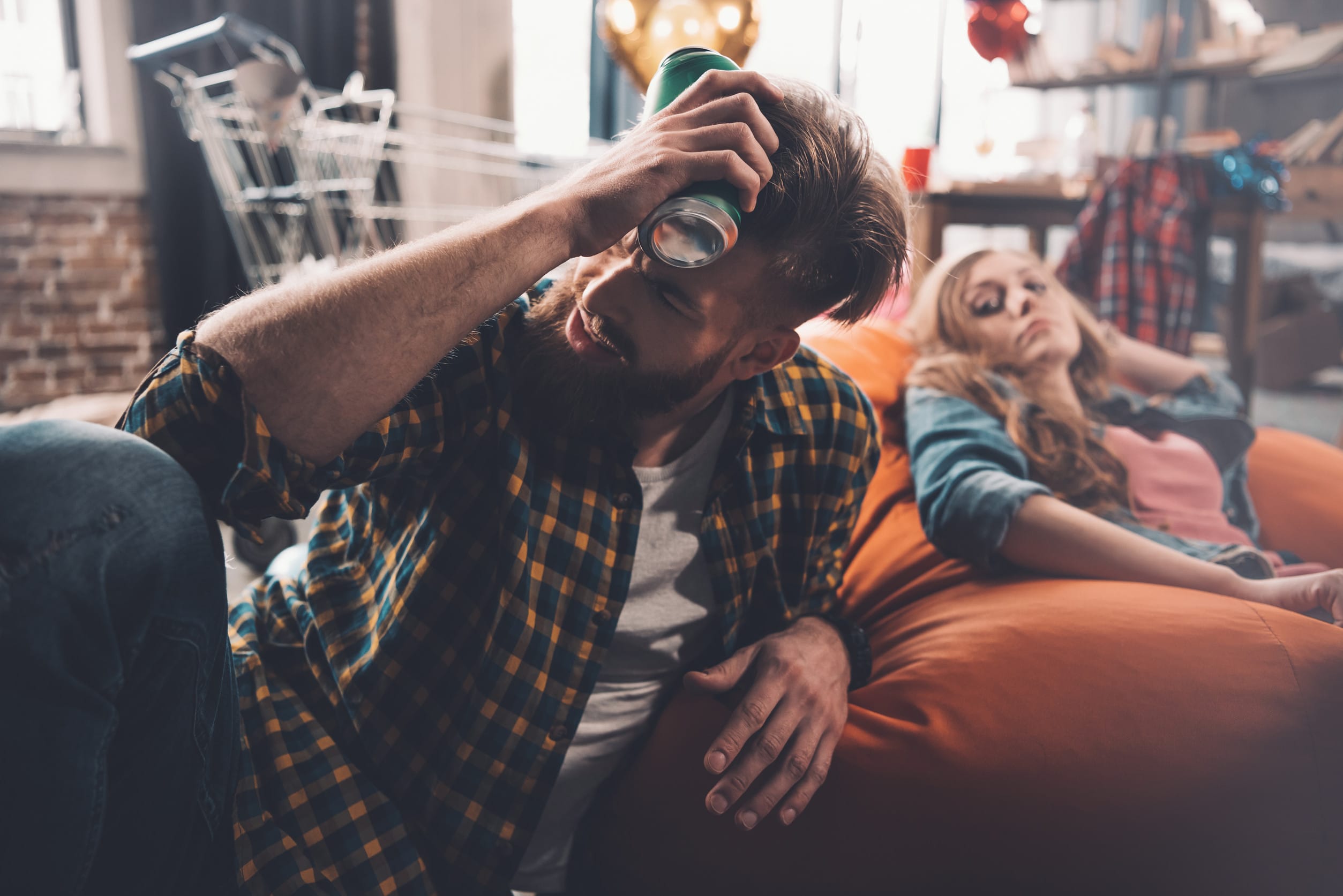 man with headache holding can of beer, woman behind in messy room after party
