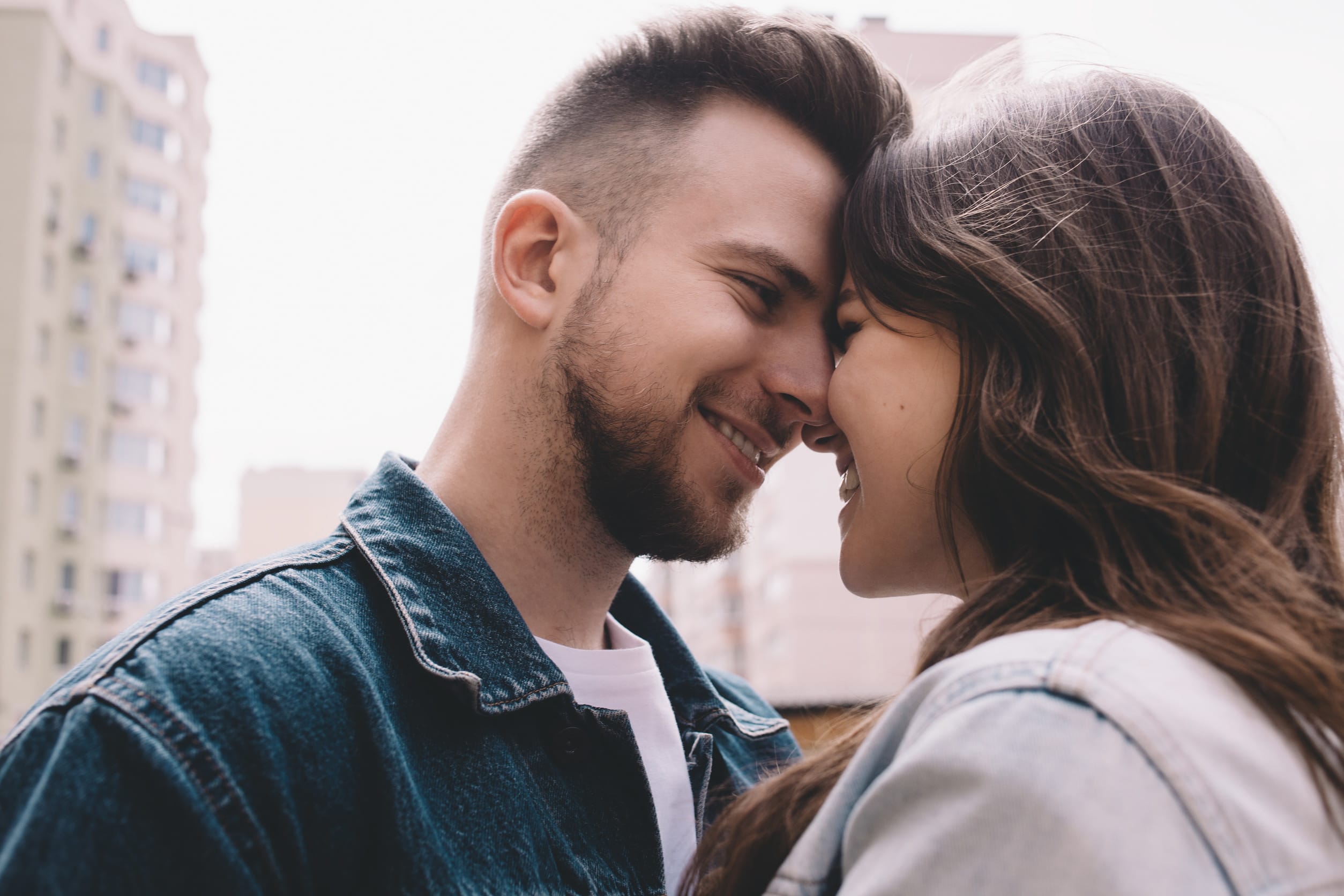 Happy attractive young couple in jeans jackets standing among buildings and trying too kiss each other. They are so delighted to have such moments. Close up