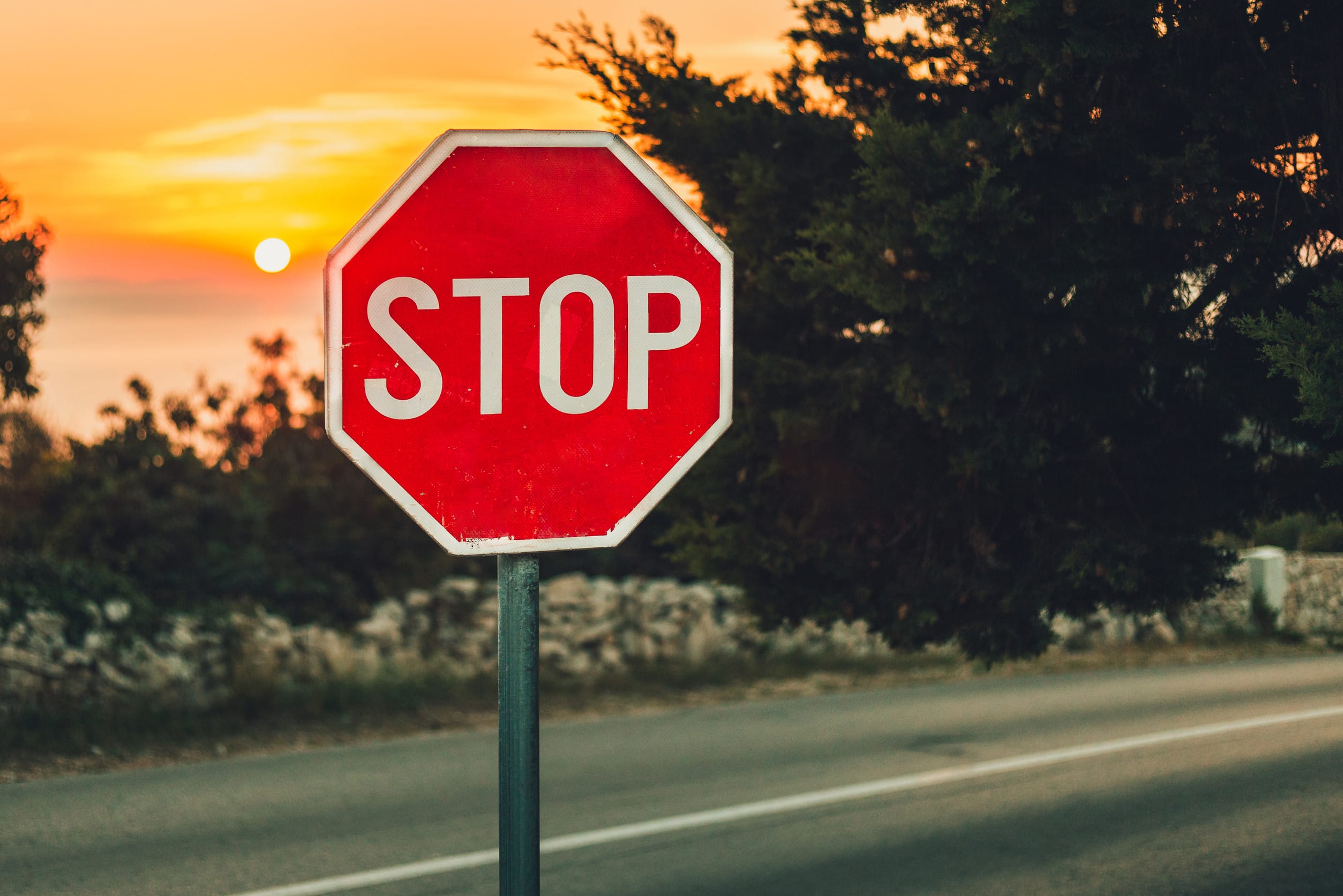 A traffic sign Stop in coastline with road and sea on background - photo on sunset. Illuminated sign by the side of the road with trees and stone wall on background. Warm toned photo of Traffic sign.