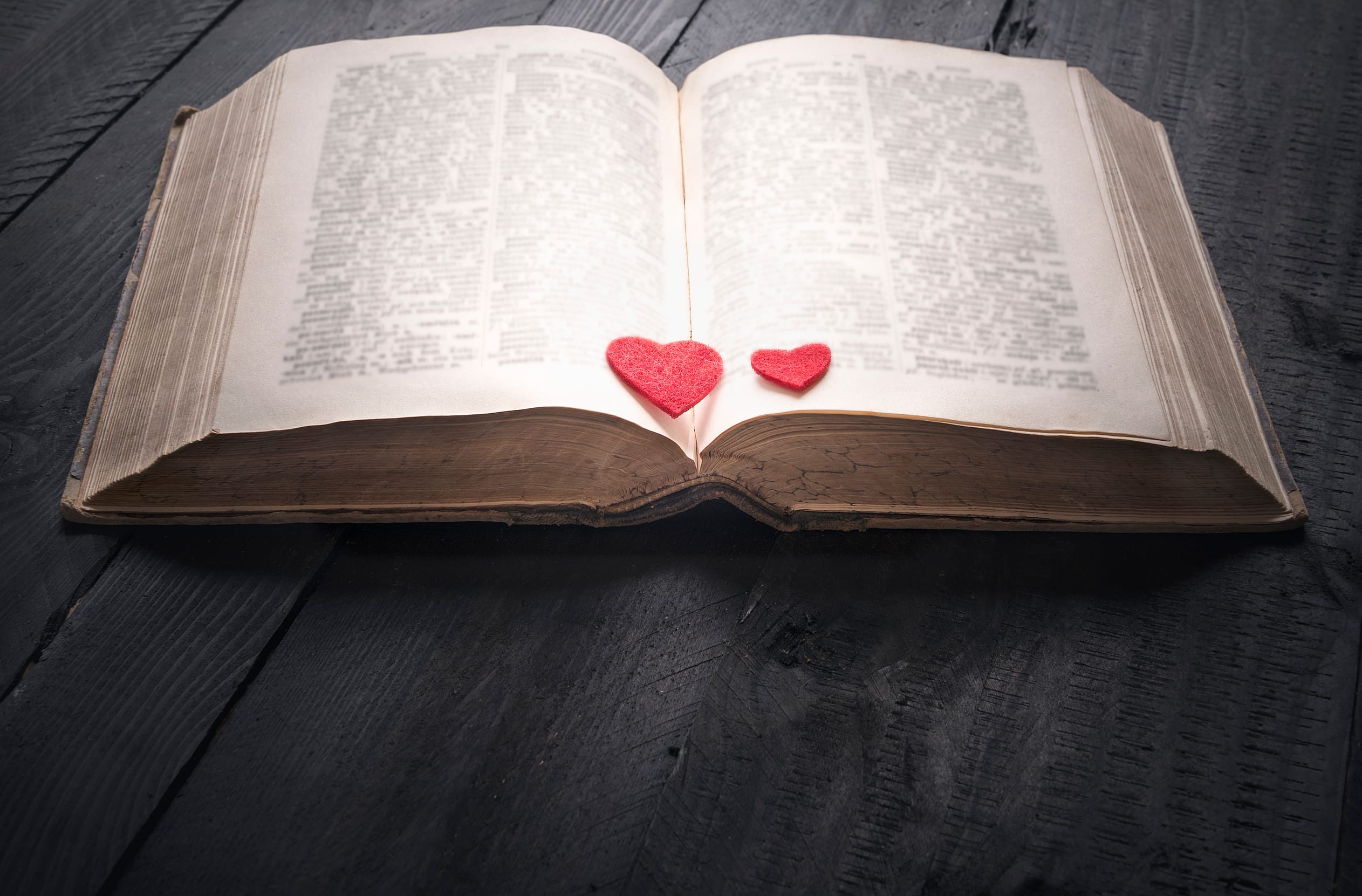 Antique open book with two red hearts on its pages, sitting on a black wooden background. A concept for the love of reading, learning, education or as a love story.