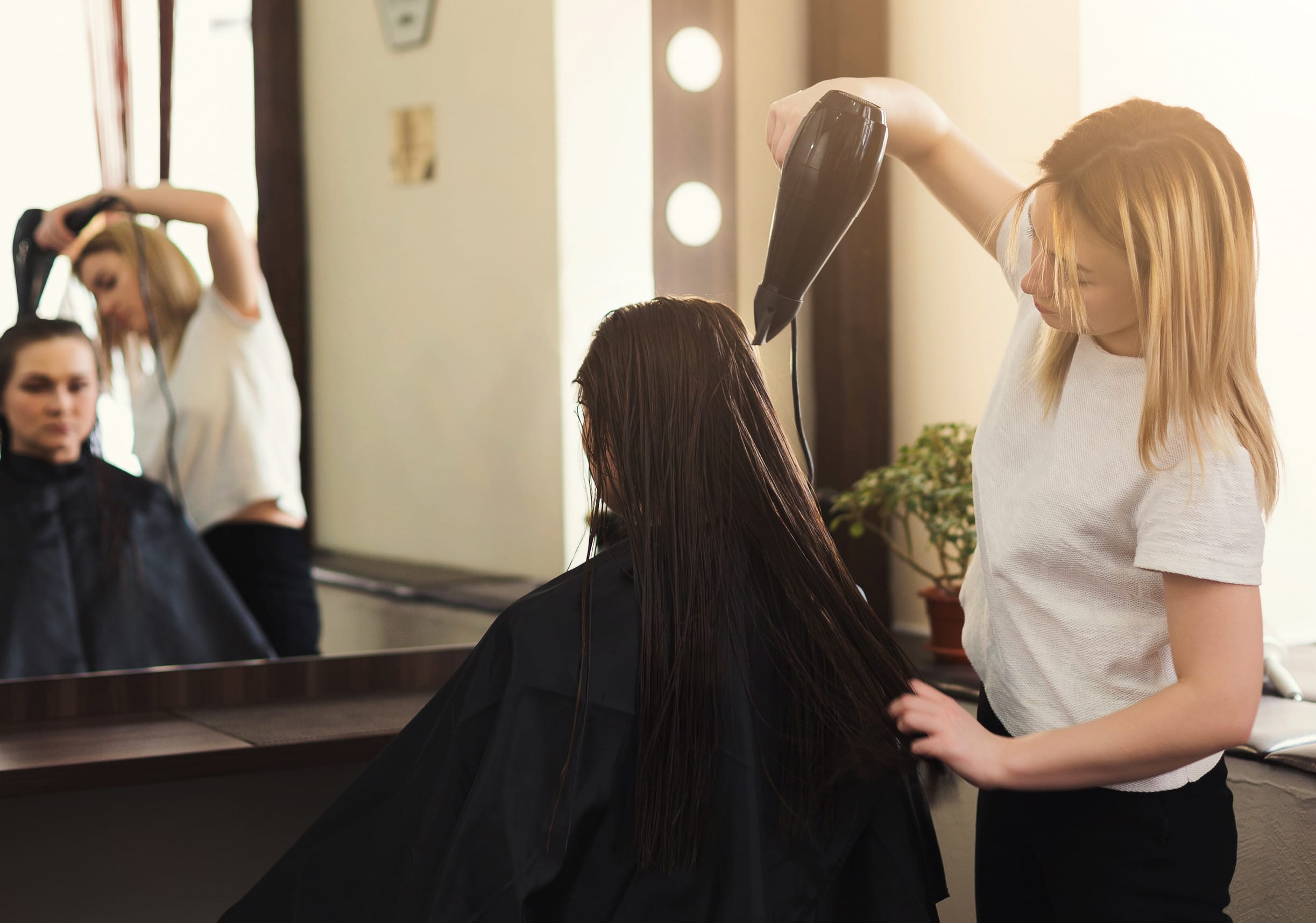 Cheerful hairdresser drying long brown hair with hair-drier in beauty salon