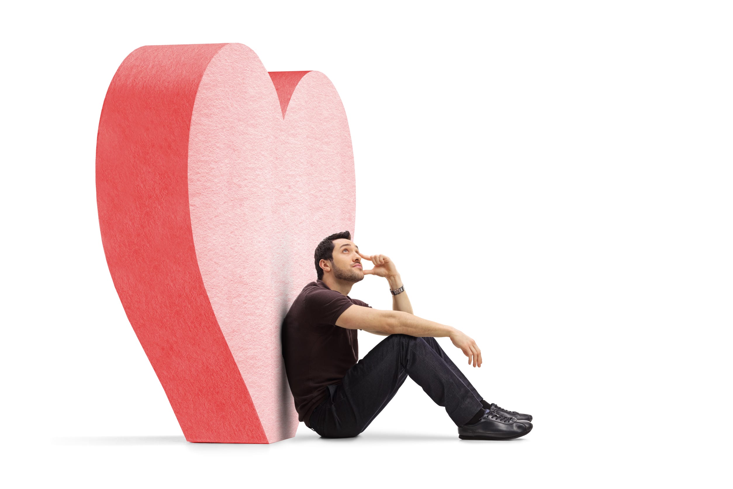 Pensive young man in love sitting next to a big red heart isolated on white background