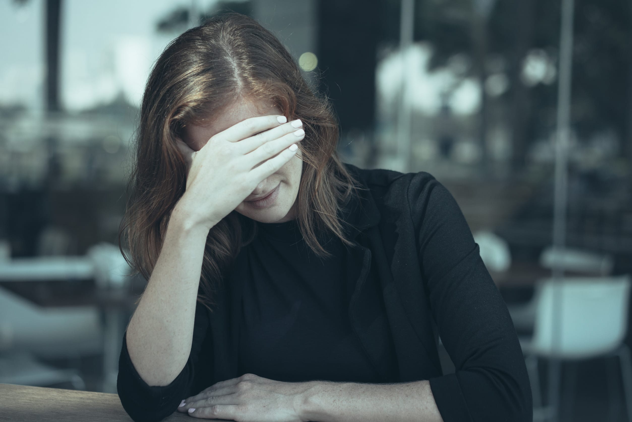 Portrait of frustrated young woman sitting at outdoor office cafe and shielding eyes with hand. Inexperienced broker after closing unprofitable deal. Work balance concept