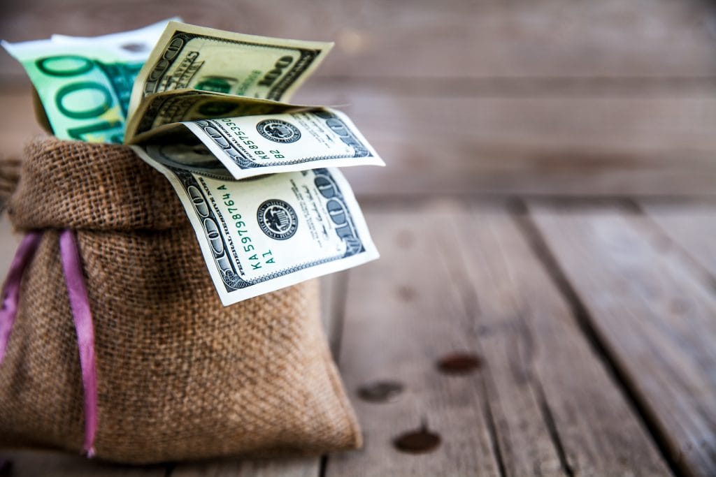 money in burlap sack with scattered money on wooden background